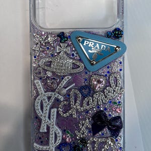 Pu&ograve; includere: Custodia trasparente per telefono con un design glitterato viola e blu. Impreziosita da strass, un logo Prada blu e altri elementi decorativi. Include un logo YSL e la parola "Chanel" in strass.