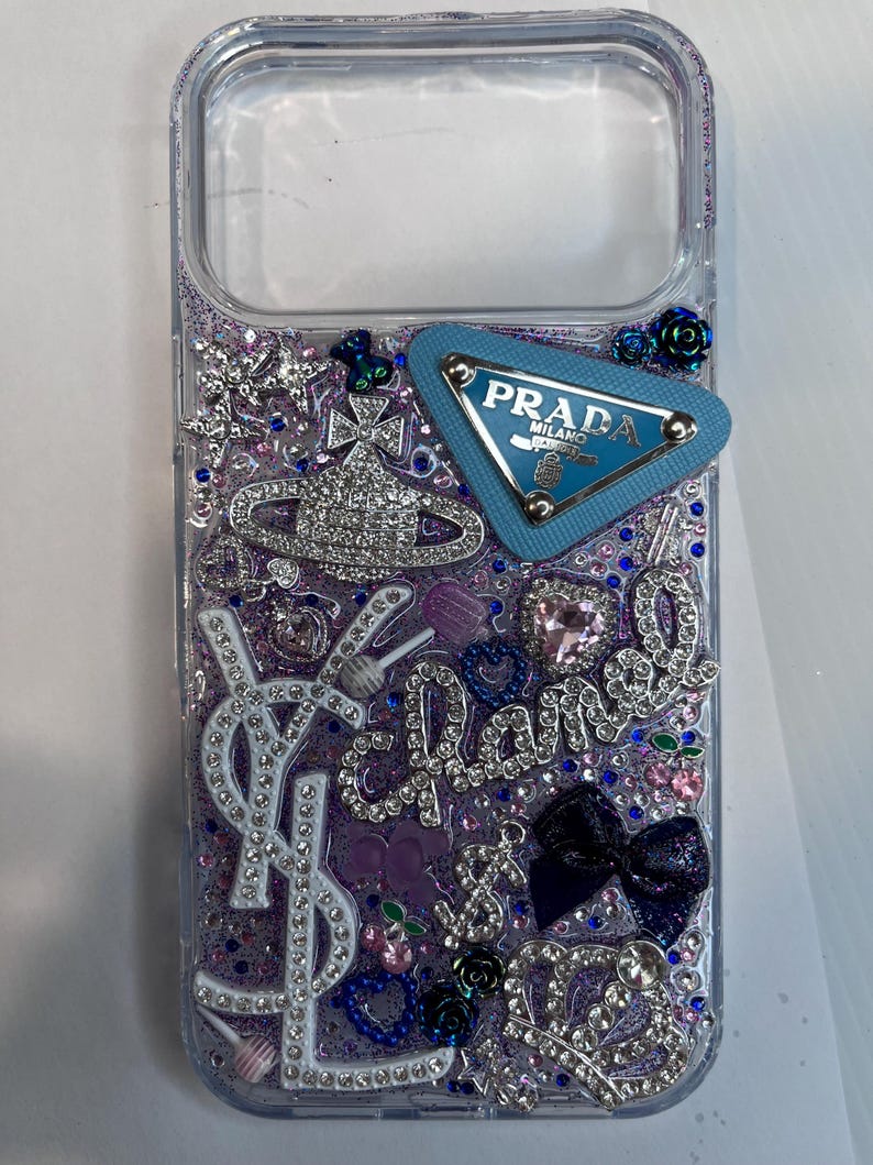 Pu&ograve; includere: Custodia trasparente per telefono con un design glitterato viola e blu. Impreziosita da strass, un logo Prada blu e altri elementi decorativi. Include un logo YSL e la parola "Chanel" in strass.