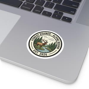 May include: A circular sticker with a nature scene featuring a brown elk standing near a river, surrounded by green trees. The text "FOREST ROAMER - ALPINE BOUND" and "EST. 2026" are in a circular design.