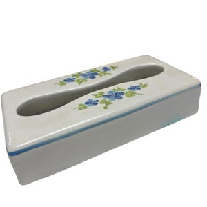 Puede incluir: Caja de pañuelos rectangular de cerámica blanca con una abertura curva. La caja está decorada con motivos florales azules y un fino borde azul. El diseño es sencillo y elegante.