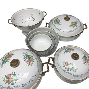 Könnte beinhalten: Eine Sammlung weißer Emaille-Küchenartikel. Enthält eine Küchensieb, Schüsseln mit Deckel und Nestschalen. Jedes Stück ist mit einem Blumenmuster in Rosa, Blau und Grün verziert, mit messingfarbenen Griffen und Akzenten. Ein Vintage-Küchenset.