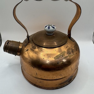 May include: A vintage copper kettle with a white and blue porcelain handle and lid knob. The kettle has a rounded body and a spout on the side. The handle is arched and attached to the body with copper brackets. The lid knob has a floral design.