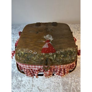 May include: A wooden picnic basket with a hinged lid featuring a hand-painted illustration of a figure in a red dress holding a pie, surrounded by white daisies. The basket is lined with a red and white chequered fabric with a ruffled edge.