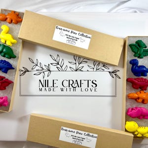 Può includere: Una collezione di pastelli a forma di dinosauro in varie tonalità di arancione, giallo, verde, blu, rosso e rosa. I pastelli sono disposti in scatole rettangolari con la scritta "Roar-some Dino Collection" e "NILE CRAFTS MADE WITH LOVE".