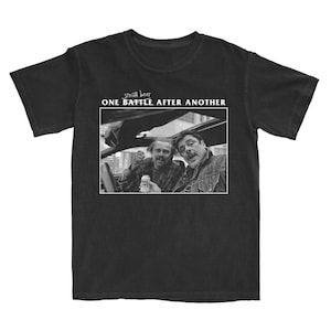 May include: Black t-shirt featuring a black and white image of two men in a car, one holding a beer. The text "small beer" is above the image, and "ONE BATTLE AFTER ANOTHER" is below it.