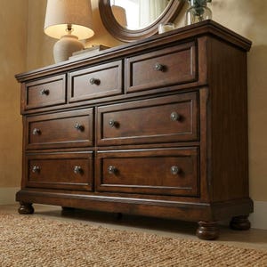 May include: A dark brown wooden dresser with seven drawers and round knobs. The dresser has a traditional design with a decorative base and is set on a woven rug. A lamp and mirror are visible in the background.