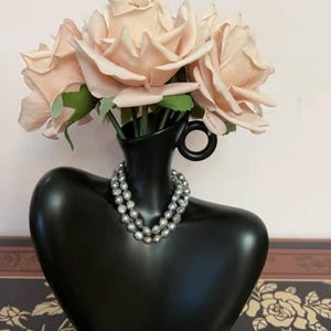 Può includere: Un vaso in ceramica nera a forma di busto, ornato da una doppia collana di perle grigie. Il vaso contiene diverse rose color pesca con foglie verdi. Il vaso ha un manico laterale.