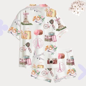 May include: White pyjama set with a repeating pattern of Parisian imagery. The design includes the Eiffel Tower, bicycles, aeroplanes, coffee cups, macarons, and luggage. The set features a short-sleeved top and shorts.