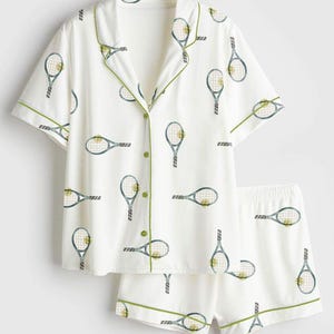 May include: White pajama set featuring a tennis racket pattern in blue and yellow. The short-sleeved top and shorts have green trim. The button-down shirt has green buttons.