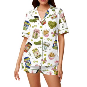 May include: White pajama set with a pickle pattern. The short-sleeved top and shorts feature a repeating design of pickle jars, pickles, and pink bows. Green trim outlines the edges of the set.