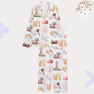May include: White pyjama set featuring a repeating pattern of Parisian travel-themed illustrations. The design includes the Eiffel Tower, cameras, bicycles, luggage, and macarons in pink, yellow, and green.