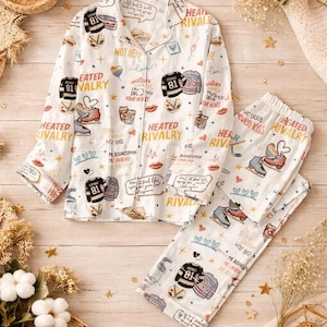 May include: White pyjama set with a button-down shirt and trousers. The set features a print of hockey jerseys, ice skates, hearts, and the words "Heated Rivalry" and "Love". The pyjamas are made of a soft material.