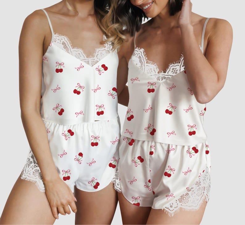 May include: White pajama set with a cherry print. The set includes a camisole top with lace trim and matching shorts. The shorts and top are decorated with red cherries and pink bows. The camisole has thin straps.