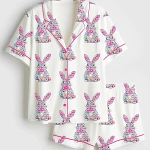 May include: White pajama set with a repeating pattern of colorful bunny illustrations. The short-sleeved top and shorts are trimmed with pink piping. The bunnies have pink bows and colorful floral patterns.