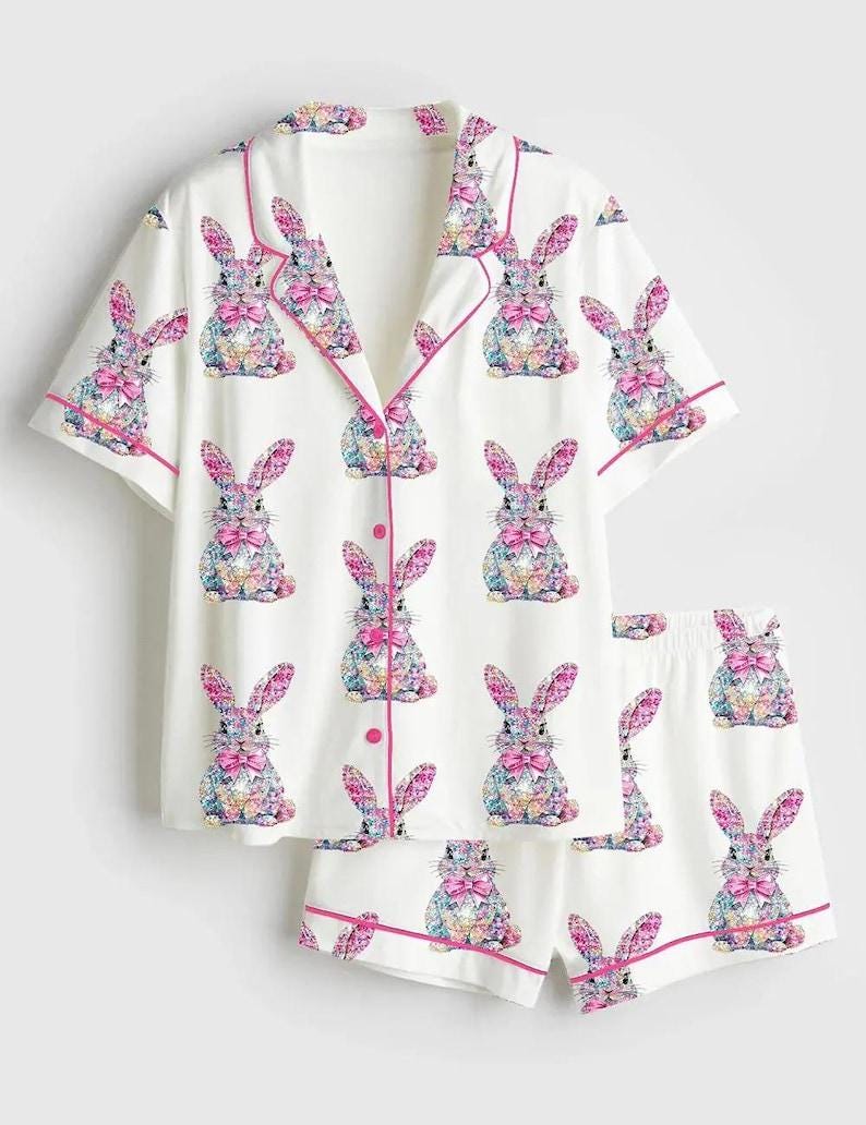 May include: White pajama set with a repeating pattern of colorful bunny illustrations. The short-sleeved top and shorts are trimmed with pink piping. The bunnies have pink bows and colorful floral patterns.