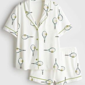 May include: White pajama set featuring a tennis racket pattern in blue and yellow. The short-sleeved top and shorts have green trim. The button-down shirt has green buttons.