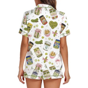 May include: White pajama set with a pickle pattern. The short-sleeved top and shorts feature illustrations of pickle jars, pickles, and pink bows. Green trim accents the edges of the set.