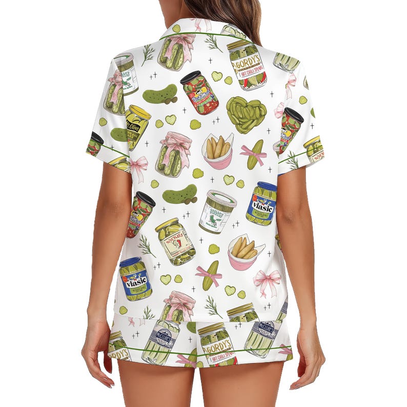 May include: White pajama set with a pickle pattern. The short-sleeved top and shorts feature illustrations of pickle jars, pickles, and pink bows. Green trim accents the edges of the set.
