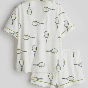 May include: White pajama set with a tennis racket pattern. The short-sleeved top and shorts feature a repeating design of tennis rackets in blue and green. Green trim accents the edges of the set, creating a sporty and playful look.