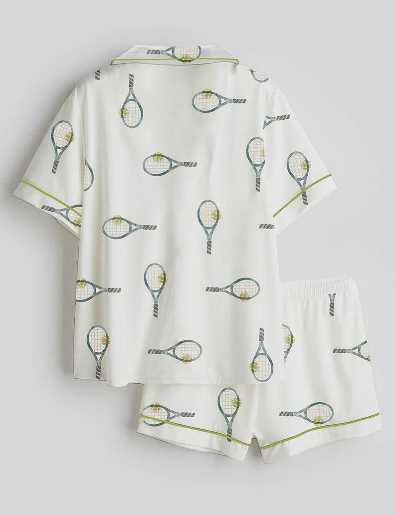 May include: White pajama set with a tennis racket pattern. The short-sleeved top and shorts feature a repeating design of tennis rackets in blue and green. Green trim accents the edges of the set, creating a sporty and playful look.