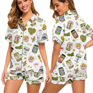 May include: White pajama set with a pickle pattern. The short-sleeved top and shorts feature illustrations of pickles, jars, and pink bows. Green trim outlines the edges of the pajamas.