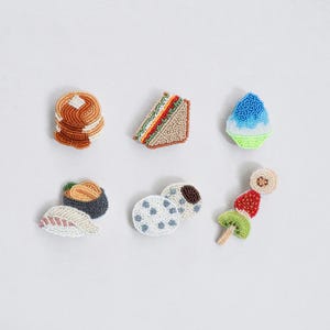 Könnte beinhalten: Eine Sammlung von Perlenbroschen mit Essensmotiven, darunter Pfannkuchen, ein Sandwich, Eis, Sushi, Kekse und ein Obstspieß. Die Broschen sind farbenfroh und detailliert, mit einer Vielzahl von Texturen und Formen.