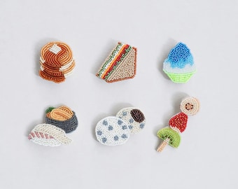 Puede incluir: Colección de broches con cuentas con temática de comida, que incluyen panqueques, un sándwich, hielo raspado, sushi, galletas y una brocheta de frutas. Los broches son coloridos y detallados, con una variedad de texturas y formas.