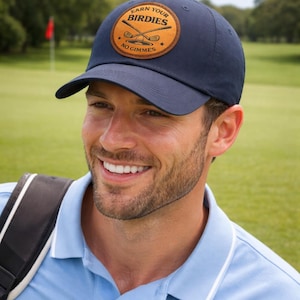 Könnte beinhalten: Marineblaue Baseballkappe mit einem runden Aufnäher mit Golfschlägern und dem Text "Earn Your Birdies, No Gimmes". Die Kappe wird von einer Person getragen, die ein hellblaues Poloshirt trägt.