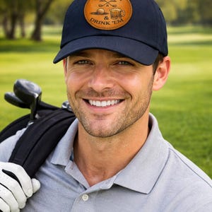 Könnte beinhalten: Marineblaue Baseballkappe mit einem braunen Lederaufnäher mit Golfmotiven und dem Text "Sink 'Em & Drink 'Em". Die Kappe wird von einer Person auf einem Golfplatz getragen.