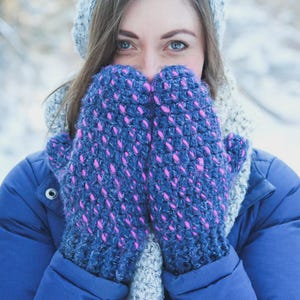May include: A person wearing a blue jacket and a gray knitted hat and scarf, holding up a pair of blue mittens with pink accents. The mittens are made of a thick, textured yarn, perfect for cold weather.