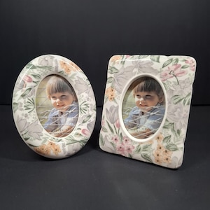 May include: Two ceramic picture frames with a floral pattern in shades of pink, green, and orange. One frame is oval and the other is rectangular with rounded corners. Both frames feature text stating "SIZE 2.5" x 3.5" MADE IN TAIWAN".