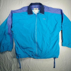 May include: A full-zip windbreaker jacket in shades of blue and purple. The jacket features a turquoise body, purple shoulders, and a navy blue collar. The jacket has a drawstring at the bottom and the brand name "NKC" is embroidered on the chest.