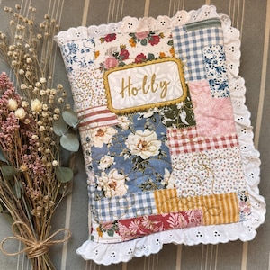 May include: A rectangular quilted fabric cover featuring a patchwork design of floral, gingham, and striped patterns in shades of blue, pink, yellow, and red. The cover has a white eyelet lace trim and a central label embroidered with the name Holly.