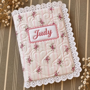 May include: A rectangular quilted fabric book cover featuring a white background with thin pink vertical stripes and small pink floral clusters. The cover has a white eyelet lace trim and a central white rectangular patch with the name Judy.
