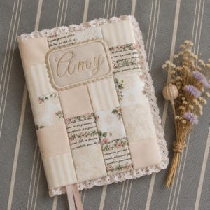 May include: A rectangular fabric-covered journal featuring a patchwork design in cream and beige tones. The cover includes floral patterns, script text, and a central embroidered patch with the name Amy. It is finished with white lace trim.