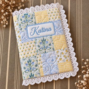 May include: A rectangular quilted fabric cover featuring a patchwork design of yellow and blue floral and gingham patterns. The center displays an embroidered white rectangular patch with the name Katina in blue script, bordered by white eyelet lace.