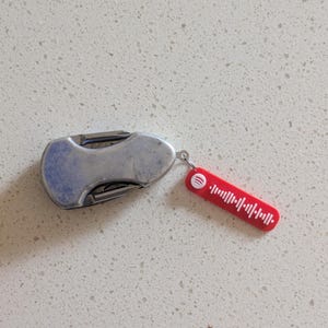 May include: A small, silver pocket knife with a curved handle and a red Spotify tag attached. The tag features a white soundwave design and the Spotify logo. The knife is closed and resting on a white speckled surface.