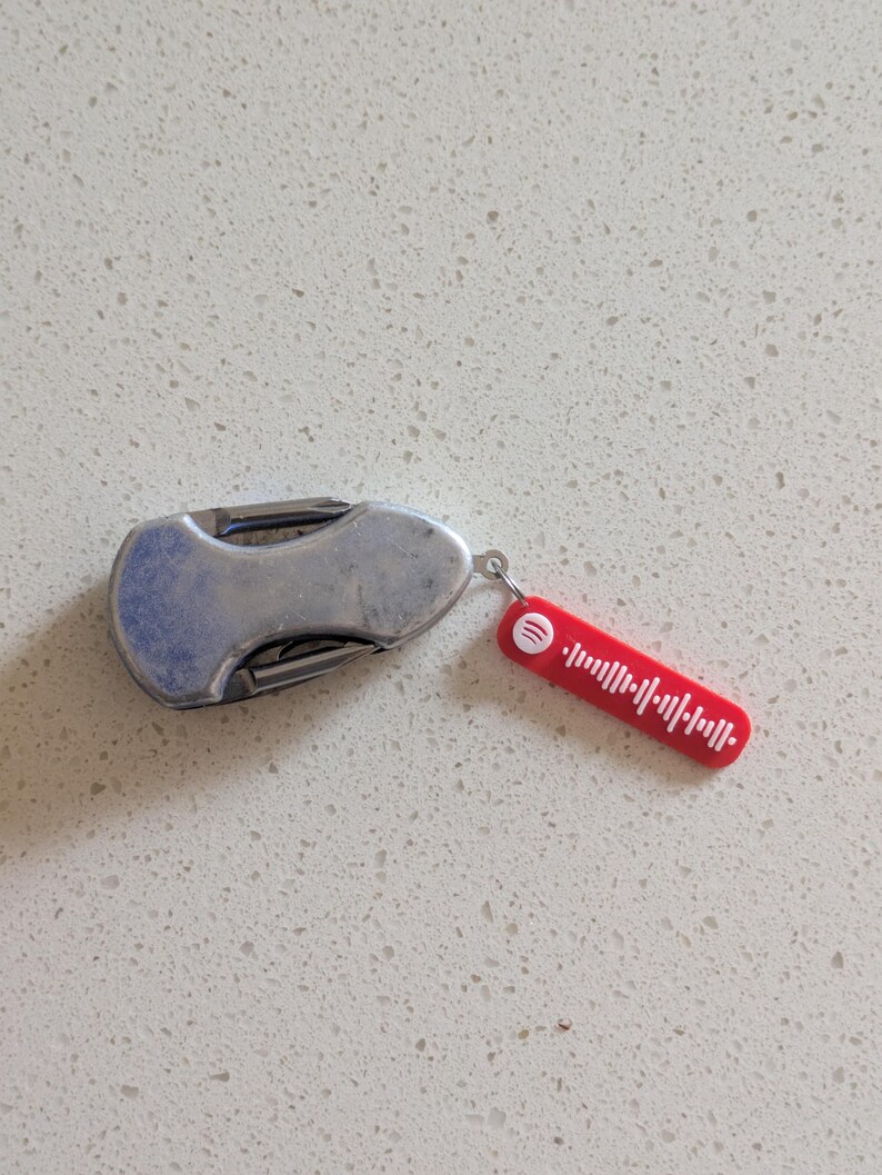 May include: A small, silver pocket knife with a curved handle and a red Spotify tag attached. The tag features a white soundwave design and the Spotify logo. The knife is closed and resting on a white speckled surface.
