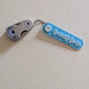 May include: A silver folding knife and a light blue Spotify keychain connected by a silver ring. The keychain has a white Spotify logo and white vertical lines representing a song's audio waveform.