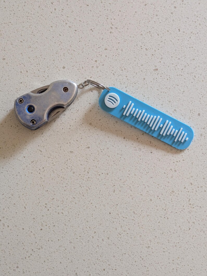 May include: A silver folding knife and a light blue Spotify keychain connected by a silver ring. The keychain has a white Spotify logo and white vertical lines representing a song's audio waveform.
