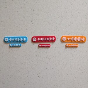 May include: Three colorful Spotify code plaques in blue, red, and orange, each with a matching smaller version. The plaques feature the Spotify logo and a series of vertical lines representing a unique audio track.
