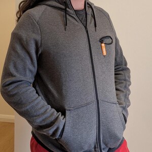 May include: A gray zip-up hoodie with a drawstring hood and two front pockets. The hoodie has a small orange tag attached to the chest. The sleeves have a textured pattern. The zipper is black.