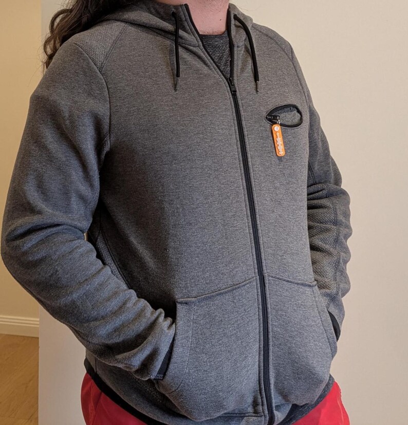 May include: A gray zip-up hoodie with a drawstring hood and two front pockets. The hoodie has a small orange tag attached to the chest. The sleeves have a textured pattern. The zipper is black.