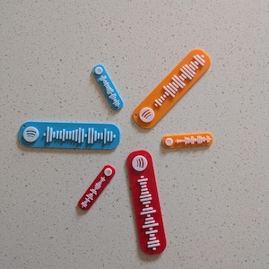 May include: A selection of colourful, rectangular Spotify code plaques in blue, orange, and red. Each plaque features a unique white waveform pattern and a Spotify logo. The plaques vary in size and are arranged on a light grey surface.