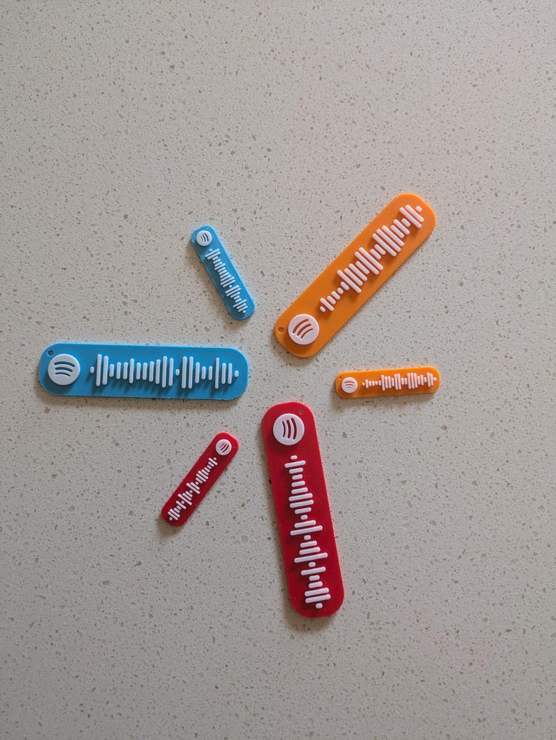May include: A collection of colorful, rectangular Spotify code plaques in blue, orange, and red. Each plaque features a unique white waveform pattern and a Spotify logo. The plaques vary in size and are arranged on a light gray surface.