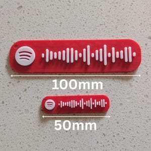 May include: Two red Spotify code tags, one 100mm long and the other 50mm long. Both tags have a white Spotify logo and a white sound wave pattern. The tags are on a light gray speckled surface.