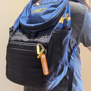 May include: A navy blue and black backpack with yellow accents, featuring the "SC" logo and "Under Armour" branding. A small orange keychain with a barcode design hangs from the zipper.