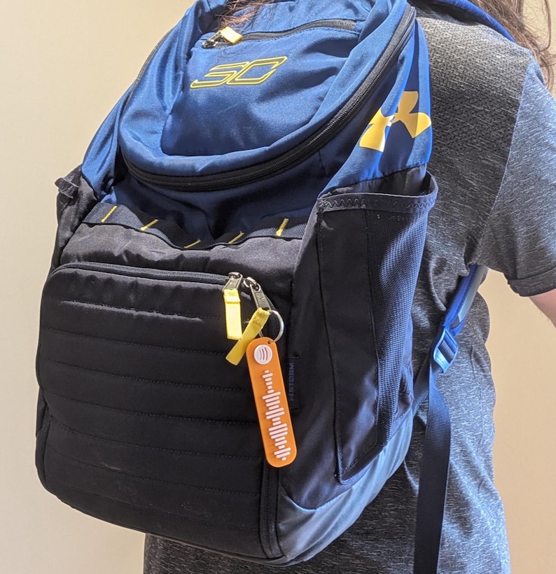 May include: A navy blue and black backpack with yellow accents, featuring the "SC" logo and "Under Armour" branding. A small orange keychain with a barcode design hangs from the zipper.