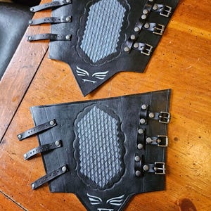 Puede incluir: Dos protectores de brazo de cuero negro con hebillas y correas plateadas. Cada protector presenta un diseño central gris texturizado dentro de un borde decorativo. Un emblema blanco en la parte inferior.