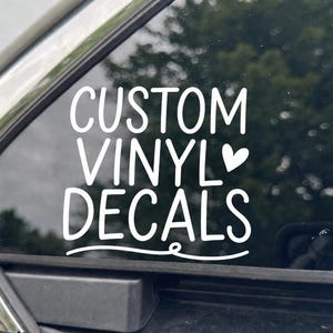 May include: A white vinyl decal applied to a car window featuring the text CUSTOM VINYL DECALS in a rounded sans-serif font. A small heart icon is positioned to the right of the word VINYL, and a decorative curved line underlines the word DECALS.